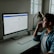 A person is sitting at a desk in front of a computer monitor displaying a financial website. The room is softly lit with natural light coming in from a nearby window. A desk lamp and a plant add to the aesthetic of the workspace. The person is holding a smartphone to their ear, appearing to be in a call.